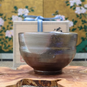 May include: A dark brown ceramic teacup with a light brown glaze. The cup is sitting on a wooden surface.