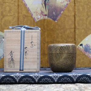May include: A wooden box with Japanese calligraphy and a blue ribbon, next to a textured, round container. The items rest on a patterned cushion, with decorative fans in the background. The background is a gold colour.