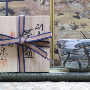 May include: A wooden box tied with a brown and blue ribbon, with Japanese calligraphy and a red stamp. Next to it is a ceramic teacup with a landscape design. A green patterned mat is in the foreground.