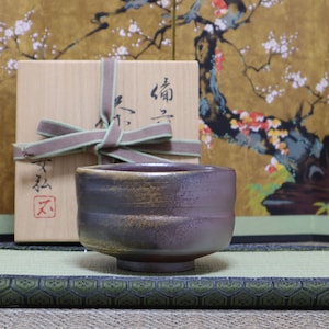 May include: A dark brown and purple ceramic tea bowl with a textured surface, sitting on a green mat with a black border. The bowl is in a wooden box with a green and brown ribbon. The box has Japanese characters on it.
