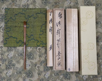 Op de afbeelding: Een bovenaanzicht van Japanse theeceremonie-artikelen. Een bruine bamboe theelepel ligt op een groen en blauw patroonmat. Daarnaast houten dozen en een cilindrische container met Japanse kalligrafie.