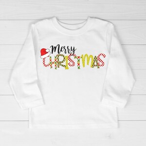 Op de afbeelding: Witte longsleeve met een rode en groene kerstthema-afbeelding met de tekst "Merry Christmas" met een kerstmanmuts op de "M".