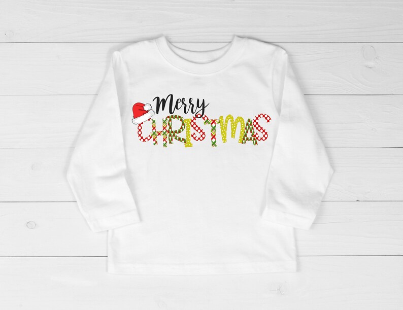 Op de afbeelding: Witte longsleeve met een rode en groene kerstthema-afbeelding met de tekst "Merry Christmas" met een kerstmanmuts op de "M".