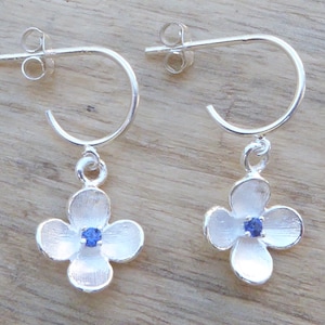 Könnte beinhalten: Silberne Creolen mit floralem Anhänger. Jede Ohrring hat einen silbernen Reifen mit einem vierblättrigen Blumenanhänger. Die Blume hat eine strukturierte Oberfläche und einen kleinen blauen Edelstein in der Mitte. Die Ohrringe werden auf einer Holzoberfläche präsentiert.