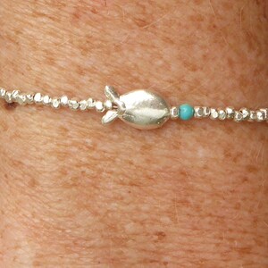 Peut inclure: Un bracelet de perles argentées avec un charm en forme de poisson et une perle turquoise. Le bracelet, porté au poignet, est composé de petites perles rondes argentées. Le charm en forme de poisson est le point central, avec la perle turquoise.
