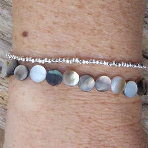May include: Two bracelets are shown. The bottom bracelet features iridescent, circular beads in shades of gray, white, and brown. The top bracelet is a delicate string of small, silver-colored beads. The bracelets are worn on a wrist.