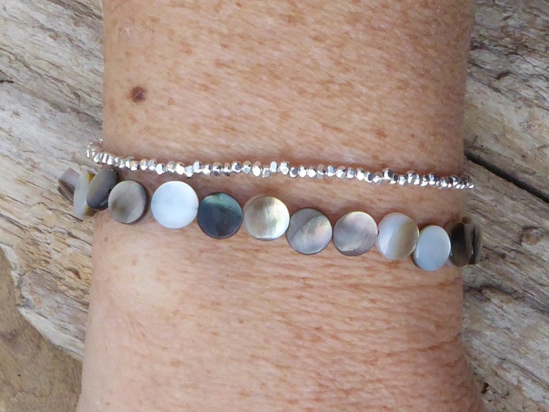 K&ouml;nnte beinhalten: Zwei Armb&auml;nder sind abgebildet. Das untere Armband hat schillernde, kreisf&ouml;rmige Perlen in Grau-, Wei&szlig;- und Braunt&ouml;nen. Das obere Armband ist eine zarte Kette aus kleinen, silberfarbenen Perlen. Die Armb&auml;nder werden an einem Handgelenk getragen.