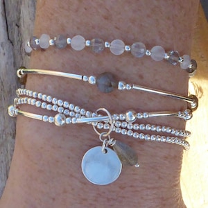 Könnte beinhalten: Ein Set aus vier silbernen Armbändern mit einem Anhänger. Die Armbänder sind an einem Handgelenk übereinander gelegt. Der Anhänger ist eine silberne Scheibe mit einer kleinen silbernen Perle daran.