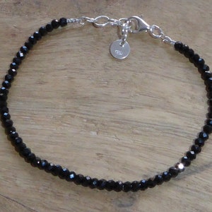 Op de afbeelding: Een delicate zwarte kralen armband met een zilveren sluiting en een klein zilveren label. De armband is gemaakt van kleine, gefacetteerde zwarte kralen.