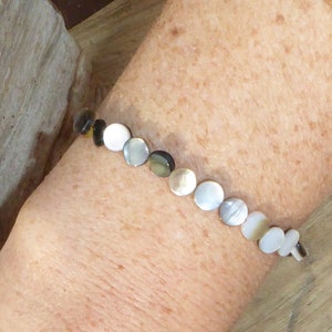 K&ouml;nnte beinhalten: Ein zartes Armband mit einer Reihe schillernder, kreisf&ouml;rmiger Perlen in Wei&szlig;-, Grau- und Braunt&ouml;nen. Das Armband wird mit einem kleinen Verschluss befestigt. Die Perlen haben einen Durchmesser von etwa 1,3 cm.