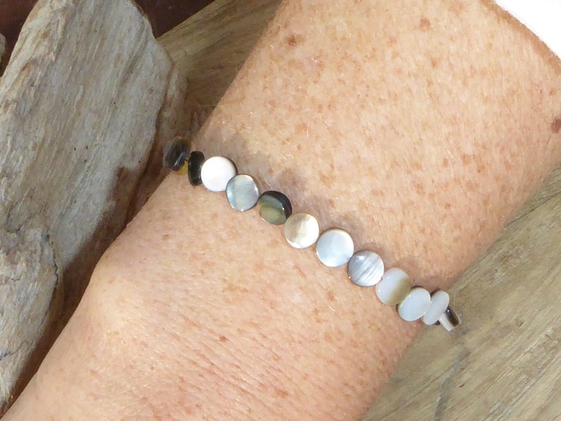 K&ouml;nnte beinhalten: Ein zartes Armband mit einer Reihe schillernder, kreisf&ouml;rmiger Perlen in Wei&szlig;-, Grau- und Braunt&ouml;nen. Das Armband wird mit einem kleinen Verschluss befestigt. Die Perlen haben einen Durchmesser von etwa 1,3 cm.