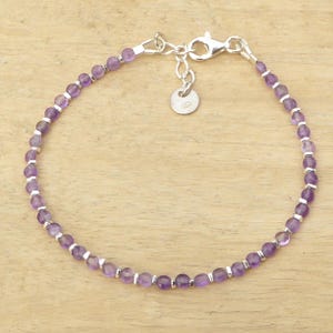 Op de afbeelding: Een delicate armband met kleine, gefacetteerde paarse kralen geregen met zilverkleurige metalen accenten. De armband heeft een karabijnsluiting en een klein, rond label. De kralen hebben een diameter van ongeveer 0,4 cm.