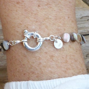 K&ouml;nnte beinhalten: Ein silbernes Armband mit schillernden, runden, muschelartigen Perlen. Das Armband hat einen gro&szlig;en, kreisf&ouml;rmigen Verschluss und einen kleinen, runden Anh&auml;nger. Die Perlen wechseln von dunklen zu hellen Farben. Das Armband wird am Handgelenk getragen.