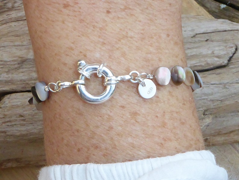 K&ouml;nnte beinhalten: Ein silbernes Armband mit schillernden, runden, muschelartigen Perlen. Das Armband hat einen gro&szlig;en, kreisf&ouml;rmigen Verschluss und einen kleinen, runden Anh&auml;nger. Die Perlen wechseln von dunklen zu hellen Farben. Das Armband wird am Handgelenk getragen.