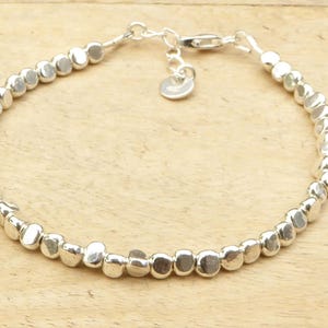 Könnte beinhalten: Ein silbernes Armband mit einer Reihe kleiner, unregelmäßiger, abgerundeter Perlen. Das Armband hat eine zarte Kette und einen Verschluss, mit einem kleinen, runden Anhänger in der Nähe des Verschlusses. Die Perlen haben einen Durchmesser von etwa 0,6 cm.