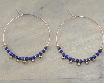 May include: Gold hoop earrings with a row of blue gemstone beads and gold beads hanging from the bottom.