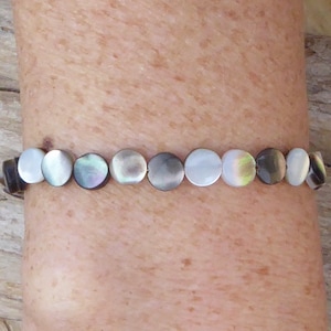 K&ouml;nnte beinhalten: Ein Armband mit schimmernden, runden Perlmuttperlen. Die Perlen sind in Wei&szlig;-, Grau- und Schwarzt&ouml;nen gehalten und auf einem dunklen Band aufgereiht. Das Armband wird an einem Arm getragen.