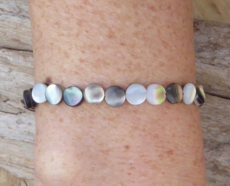 K&ouml;nnte beinhalten: Ein Armband mit schimmernden, runden Perlmuttperlen. Die Perlen sind in Wei&szlig;-, Grau- und Schwarzt&ouml;nen gehalten und auf einem dunklen Band aufgereiht. Das Armband wird an einem Arm getragen.