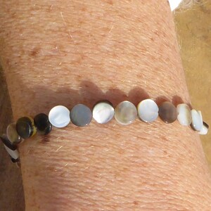 K&ouml;nnte beinhalten: Ein Armband aus runden, irisierenden Perlmuttperlen. Die Perlen sind in Wei&szlig;-, Grau-, Braun- und Schwarzt&ouml;nen gehalten. Das Armband wird am Handgelenk getragen.