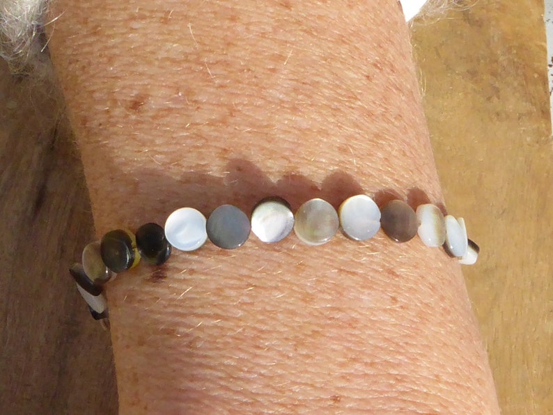 K&ouml;nnte beinhalten: Ein Armband aus runden, irisierenden Perlmuttperlen. Die Perlen sind in Wei&szlig;-, Grau-, Braun- und Schwarzt&ouml;nen gehalten. Das Armband wird am Handgelenk getragen.