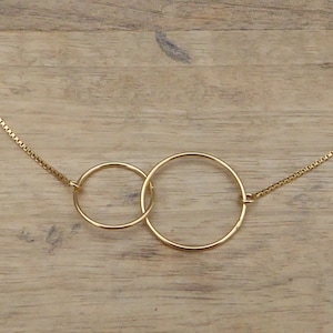 May include: A gold-colored necklace featuring two interlocking circles. The circles are connected by a delicate chain. The necklace is displayed on a light brown wooden surface.
