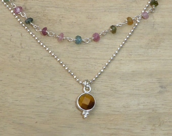 May include: A silver chain necklace with a round brown tiger's eye gemstone pendant. The necklace has a second, shorter chain with small pink, green, and blue gemstones.