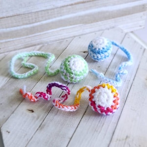 May include: A collection of colorful, handmade crochet cat toys. The toys include balls and spiral shapes in white, green, blue, orange, purple, and yellow. The toys are arranged on a white wooden surface.