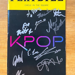 Puede incluir: Un programa de mano con una sección superior amarilla y una sección inferior negra. La palabra "PLAYBILL" está en letras blancas en negrita. La palabra "KPOP" está en rosa y azul. El programa de mano está cubierto de firmas.