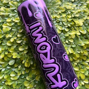 Pode incluir: Um copo de a&ccedil;o inoxid&aacute;vel roxo e preto com um design roxo que pinga e a palavra "Amanh&atilde;" escrita em uma fonte de bolhas roxa. O copo tem um fundo preto com um padr&atilde;o de pequenos cora&ccedil;&otilde;es.