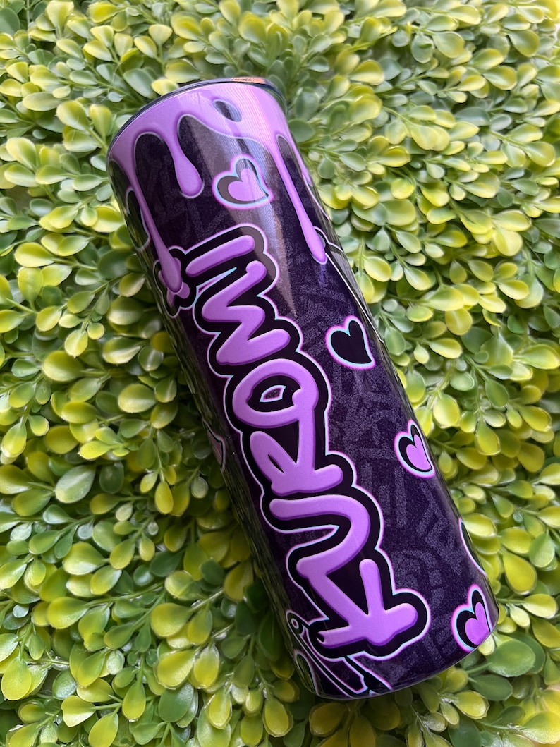 Pode incluir: Um copo de a&ccedil;o inoxid&aacute;vel roxo e preto com um design roxo que pinga e a palavra "Amanh&atilde;" escrita em uma fonte de bolhas roxa. O copo tem um fundo preto com um padr&atilde;o de pequenos cora&ccedil;&otilde;es.