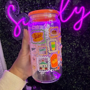 May include: A clear glass jar with an orange lid, decorated with colorful stickers. The stickers feature various designs, including a coffee cup, pancakes, and the text "Luke's Gilmore Girls". The jar is held in a hand.