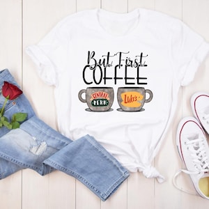 May include: White t-shirt with the phrase "But First Coffee" in black script. The shirt features two coffee cup designs, one with "Central Perk" and the other with "Luke's". The image includes blue jeans, a red rose, and white sneakers.