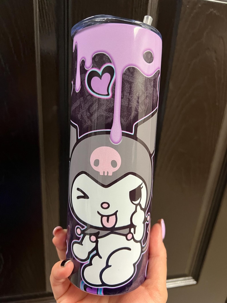 Pode incluir: Um copo de a&ccedil;o inoxid&aacute;vel com um personagem de desenho animado preto e branco com uma caveira e ossos cruzados na cabe&ccedil;a. O copo tem um design de gotejamento roxo e um cora&ccedil;&atilde;o rosa.