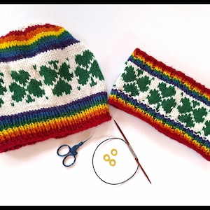 Op de afbeelding: Een witte gebreide muts en bijpassende hoofdband met een regenboogstreep en een groen klaverbladpatroon.