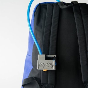 May include: A gray plastic "Sip Clip" attached to a black and blue backpack. A blue hydration tube is held by the clip. The text "Sip Clip" is printed on the clip.