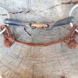 May include: A horse bit featuring a dark metal mouthpiece and large silver rings. Brown rope knots and a braided section connect the bit. The bit is displayed on a weathered, light brown wooden surface.