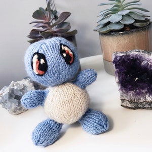 May include: A blue and cream knitted turtle toy with embroidered eyes. The toy is sitting on a white surface with a purple amethyst crystal and a succulent plant in a brown pot.