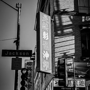 May include: A monochrome street scene featuring a variety of signs in English and Chinese. The image includes a sign for "Jackson", traffic lights, and various business signs. The photograph was taken during the day.