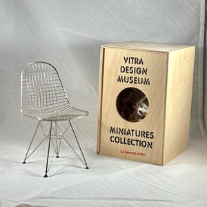 May include: A miniature wire chair with a silver finish stands next to a wooden box. The box features the text "Vitra Design Museum Miniatures Collection" and a circular opening. The chair has a geometric design.
