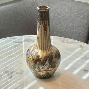 May include: A ceramic vase with a tall, slender neck and a rounded base, featuring a glossy finish in shades of cream, brown, and muted green. The surface is decorated with a repeating pattern of stylized dragonfly motifs and vertical ribbed textures.
