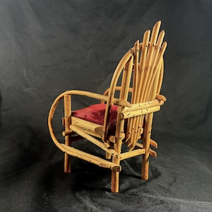May include: A miniature wooden chair with a rustic design. The chair features a red velvet seat and a backrest made of vertical wooden rods. The frame is constructed from natural-colored branches, showcasing a handcrafted aesthetic. The chair is a decorative item.