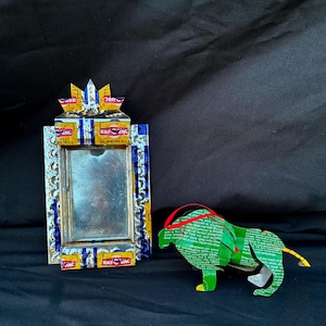 May include: A handcrafted mirror and lion sculpture. The mirror frame is made from recycled materials with blue, yellow, and silver details. The lion is green with a red headband and yellow tail, also made from recycled materials.