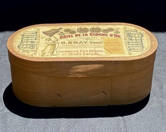 May include: A vintage wooden box with an oval lid. The lid features a printed label with the text "HOTEL DE LA CLOCHE D'OR" and illustrations. The box is made of light-colored wood and has a simple, classic design.