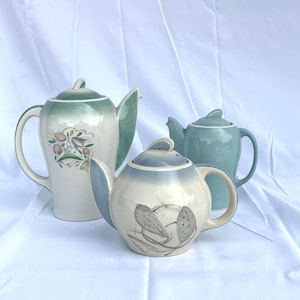 May include: Three ceramic teapots in shades of green and blue. The teapots have curved handles, spouts, and lids. One teapot has a floral design, while another features a feather motif. The teapots are set against a white background.