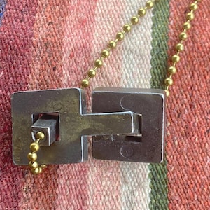 May include: A silver-coloured pendant necklace with a geometric design. The pendant features two square shapes connected by a rectangular bar. The necklace has a gold-coloured ball chain. The background is a colourful, textured fabric.