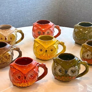 May include: A set of ceramic owl-shaped mugs in shades of orange, yellow, brown, and green. Each mug features a handle and a detailed owl face design. The mugs are displayed on a white, round table.