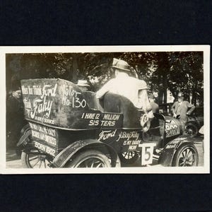 May include: A black and white photo of a vintage Ford Model T car with the text "Think Ford Motor Fahy No. 130" and "I have 12 million sis ters" on the side. The car is parked on a street and there are people standing around it.