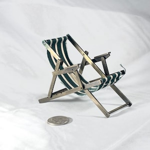 May include: A small, folding beach chair made of silver-toned metal and green and white striped fabric. The chair is shown folded. A coin is next to the chair for scale. The chair's dimensions are approximately 10 cm.