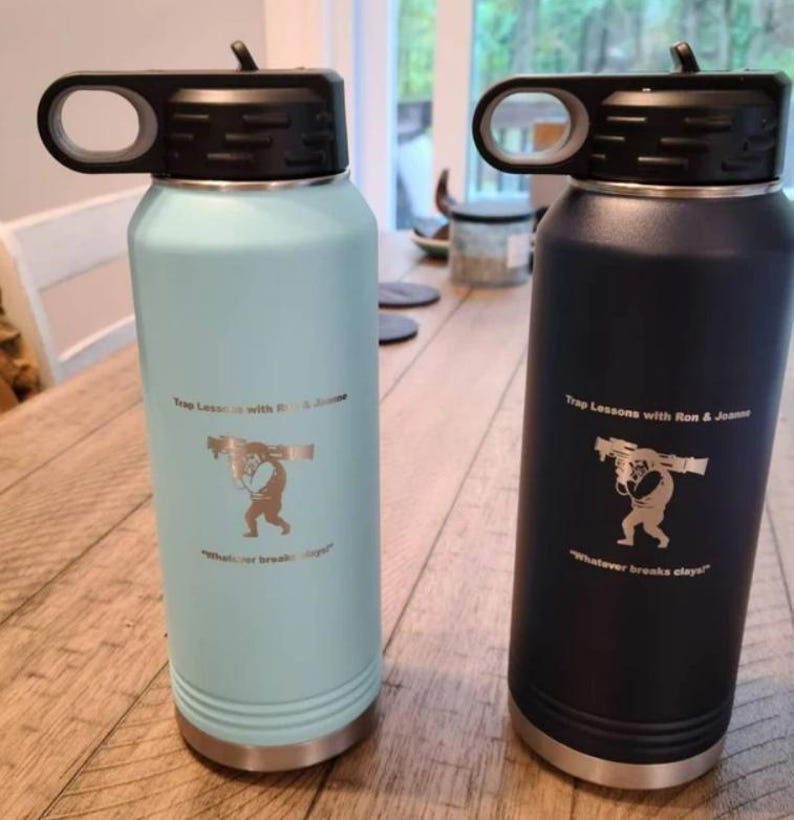 May include: Two stainless steel water bottles with black lids and handles. The bottle on the left is light blue and the bottle on the right is dark blue. Both bottles have a graphic of a person carrying a log on their back and the text "Trap Lessons with Ron & Joanne" and "Whatever breaks clays".