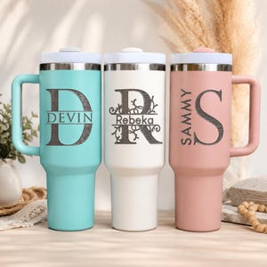 May include: Three insulated tumblers with handles and lids. The left is teal with a large letter D and the name DEVIN. The center is white with a large letter R and the name Rebeka. The right is dusty pink with a large letter S and the name SAMMY.
